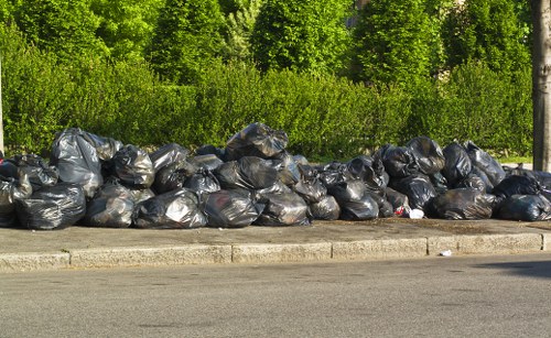 A long row of black trash bags filled with rubbish is lined up along the edge of a paved street or driveway in an outdoor setting, with lush green trees and bushes in the background. The bags appear to contain general waste and are stacked closely together, resting on the curb adjacent to the pavement. The leak-proof plastic bags exhibit a shiny surface, and some are slightly bulging, indicating they are filled to capacity. The scene suggests a waste collection or removal activity, possibly handled by Commercial Waste Removal West Ham, with the bags positioned for pickup or disposal. The environment appears calm and residential or semi-urban, with no other objects or vehicles visible, highlighting a typical waste management process in a local area near West Ham, with the scene captured in clear daylight.
