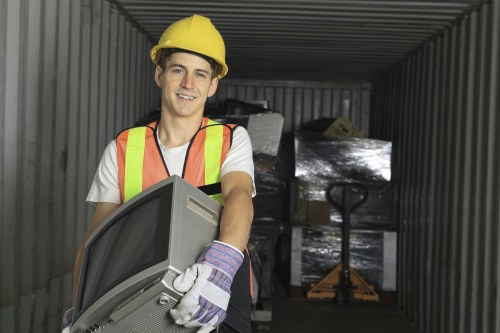 A young male waste management worker wearing a yellow safety helmet, high-visibility vest, and work gloves is standing inside a metal storage container or shipping skip. He is holding a computer monitor, suggesting involvement in administrative or logistical tasks related to waste collection. Behind him, there are stacked cardboard boxes wrapped in plastic, located on a wooden pallet, which appear to be part of waste or recycled material. The container's interior has corrugated metal walls, and a manual pallet truck is visible in the background, indicating a waste storage or disposal area. The lighting is natural, illuminating the worker's face and the surrounding objects clearly. This scene reflects a professional rubbish removal environment consistent with services such as those offered by Commercial Waste Removal West Ham, supporting efficient waste management and disposal operations in the local area around the postcode for West Ham.