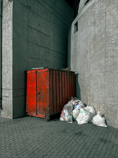 A black cylindrical rubbish bin with a partially open white plastic lid is positioned against a large, smooth stone wall in an indoor or outdoor public space. The top of the bin is cluttered with vari