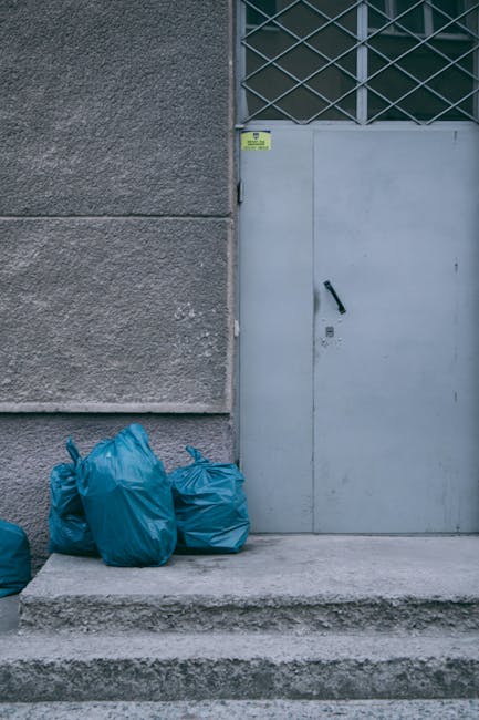 In the foreground, there are three blue garbage bags made of synthetic material placed on a concrete doorstep outside a building. The bags appear to be filled with waste and are tied at the top. The b
