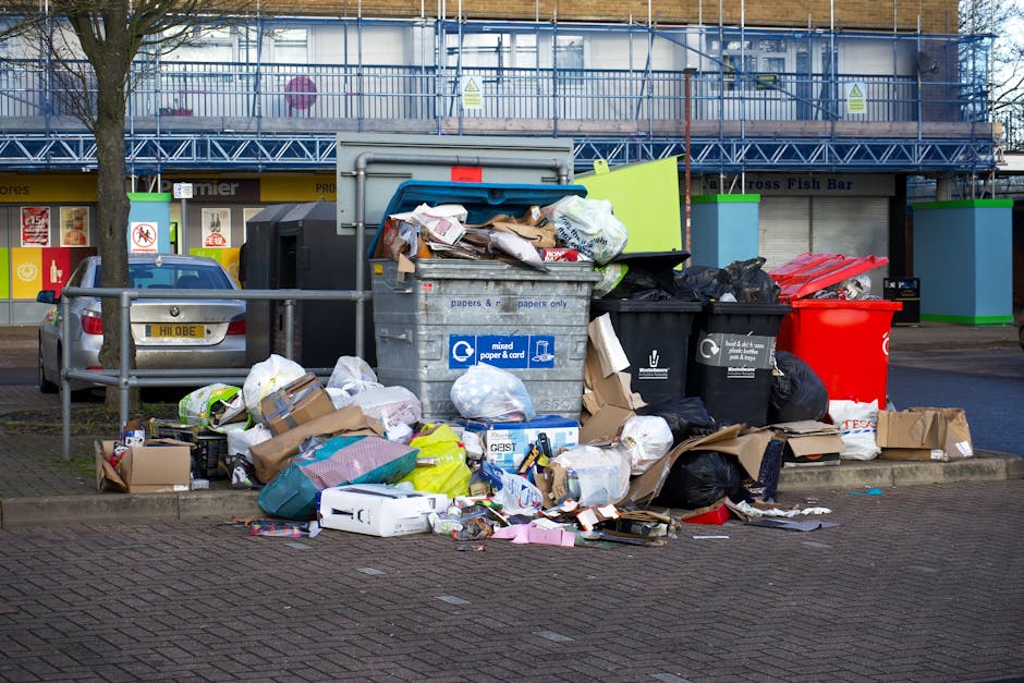 A middle-aged man with dark hair, dressed in a black t-shirt and dark trousers, is bending over a stainless steel outdoor rubbish bin to dispose of waste. He is holding a large white garbage bag fille